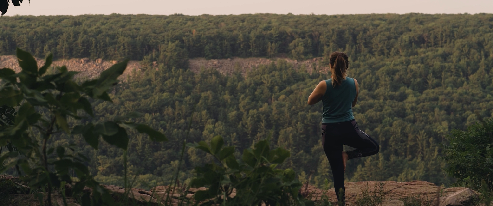 Aktiv Style USA cinematic brand commercial still, a woman in a yoga tree pose on a bluff overlooking a forested valley